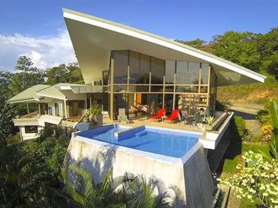 White hillside house beside a swimming pool, surrounded by tropical trees.