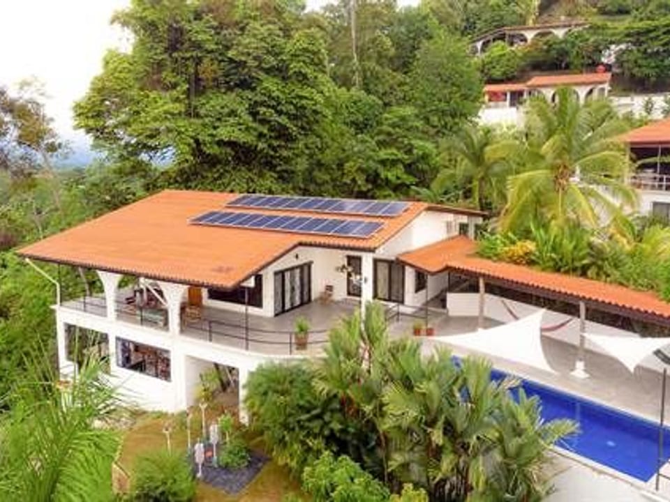 House with rooftop solar panels and a pool among dense tropical greenery.