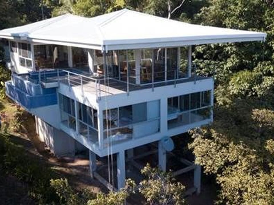 Elevated contemporary house with wide windows overlooking tropical trees.